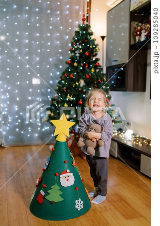 Little girl squints and grimaces while standing with a teddy bear near a decorated toy Christmas tree 109285040