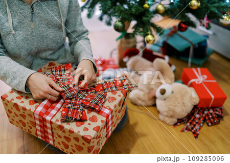 Girl unties a bow on a colorful gift box sitting near the Christmas tree. Cropped. Faceless 109285096