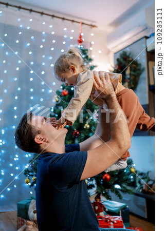 Dad lifts a little girl up while sitting near a sparkling Christmas tree. Side view 109285111