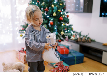 Little girl holds a toy duck in her hands while standing on a decorated Christmas tree 109285123