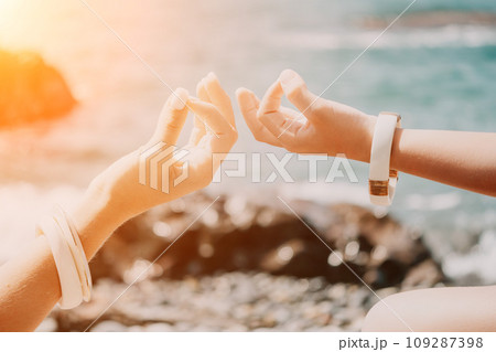 Close up Yoga Hand Gesture of Woman Doing an Outdoor meditation. Blurred sea background. Woman on yoga mat in beach meditation, mental health training or mind wellness by ocean, sea Close up Yoga Hand Gesture of Woman Doing an Outdoor meditation. Blurred sea background. Woman on yoga mat in beach meditation, mental health training or mind wellness by ocean, sea 109287398