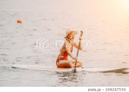 Woman in red bikini on sup board. Happy lady with blond hair in red bathing suit chilling and sunbathing by turquoise sea ocean on sunset. Holiday, vacation and recreational concept. Woman in red bikini on sup board. Happy lady with blond hair in red bathing suit chilling and sunbathing by turquoise sea ocean on sunset. Holiday, vacation and recreational concept. 109287550