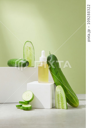The serum bottle containing light yellow liquid is placed on a white cube-shaped platform, surrounded by cucumber decoration. Vegan cosmetics with cucumber extract. The serum bottle containing light yellow liquid is placed on a white cube-shaped platform, surrounded by cucumber decoration. Vegan cosmetics with cucumber extract. 109288322