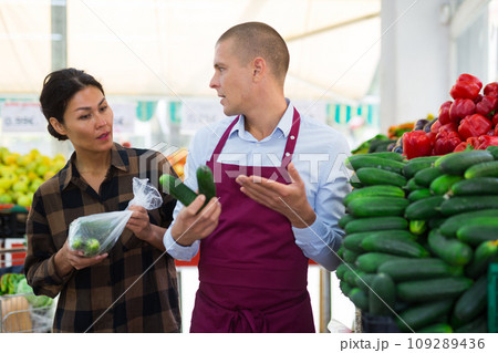 Greengrocer worker helping woman to choose cucumbers Greengrocer worker helping woman to choose cucumbers 109289436