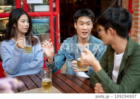 A happy young Asian man is sharing his funny story while enjoying beers with his friends at a bar. 109294887