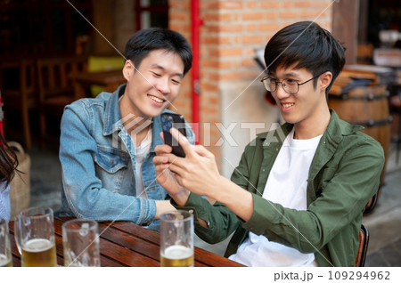 Two happy Asian male friends are watching a funny video on a phone while hanging out at a restaurant 109294962