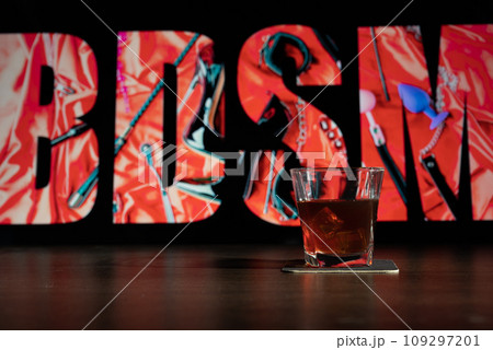 Glass of whiskey on the bar in front of the blur image Creative BDSM lettering on a black background. Set of sexy erotic toys Glass of whiskey on the bar in front of the blur image Creative BDSM lettering on a black background. Set of sexy erotic toys 109297201