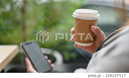 Closeup young woman holding paper cup with coffee and using smartphone at cafe Closeup young woman holding paper cup with coffee and using smartphone at cafe 109297517