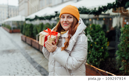 Happy young beautiful excited woman holding gift box in hands and smile on winter street. Winter holidays sale. Concept of shopping, holidays and happiness christmas. High quality photo 109297914