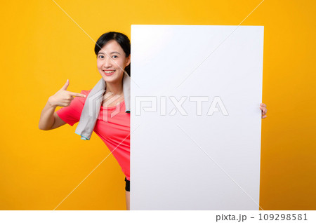 young asian sports fitness woman happy smile wearing pink sportswear standing behind the white blank banner or empty space advertisement board against yellow background. wellbeing lifestyle concept. young asian sports fitness woman happy smile wearing pink sportswear standing behind the white blank banner or empty space advertisement board against yellow background. wellbeing lifestyle concept. 109298581