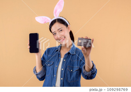 Portrait young asian woman wearing denim clothes and bunny rabbit ears and holding smartphone and credit card isolated on beige background. Shopping Happy Easter concept. Portrait young asian woman wearing denim clothes and bunny rabbit ears and holding smartphone and credit card isolated on beige background. Shopping Happy Easter concept. 109298769