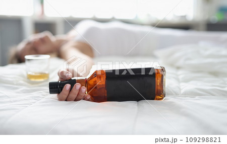 Woman lies in white bed with bottle of whiskey in hand 109298821