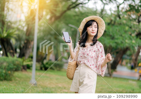 Portrait of asian young woman traveler with weaving hat and basket using mobile phone on green park nature background. Journey trip lifestyle, world travel explorer or Asia summer tourism concept. 109298990