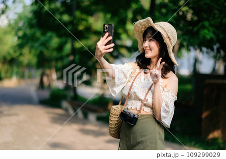 Portrait of asian young woman traveler with weaving hat, basket, mobile phone and camera on green public park background. Journey trip lifestyle, world travel explorer or Asia summer tourism concept. Portrait of asian young woman traveler with weaving hat, basket, mobile phone and camera on green public park background. Journey trip lifestyle, world travel explorer or Asia summer tourism concept. 109299029