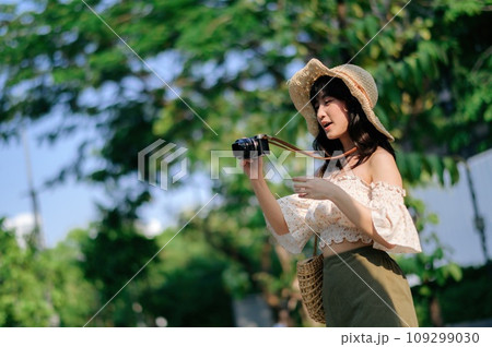 Portrait of asian young woman traveler with weaving hat and basket and a camera on green public park nature background. Journey trip lifestyle, world travel explorer or Asia summer tourism concept. Portrait of asian young woman traveler with weaving hat and basket and a camera on green public park nature background. Journey trip lifestyle, world travel explorer or Asia summer tourism concept. 109299030