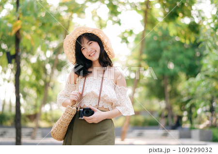 Portrait of asian young woman traveler with weaving hat and basket and a camera on green public park nature background. Journey trip lifestyle, world travel explorer or Asia summer tourism concept. Portrait of asian young woman traveler with weaving hat and basket and a camera on green public park nature background. Journey trip lifestyle, world travel explorer or Asia summer tourism concept. 109299032