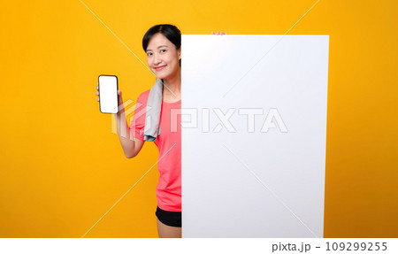 young asian sports fitness woman happy smile wearing pink sportswear standing behind the white blank banner or empty space advertisement board against yellow background. wellbeing application concept. young asian sports fitness woman happy smile wearing pink sportswear standing behind the white blank banner or empty space advertisement board against yellow background. wellbeing application concept. 109299255