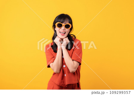 Portrait young beautiful asian woman cute and shy dressed in orange clothes and sunglasses touching cheeks showing shy emotional feeling isolated on yellow studio background. Portrait young beautiful asian woman cute and shy dressed in orange clothes and sunglasses touching cheeks showing shy emotional feeling isolated on yellow studio background. 109299357