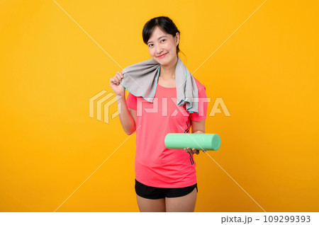 Portrait young asian sports fitness woman happy smile wearing pink sportswear and face towel doing exercise training workout against yellow studio background. wellbeing and healthy lifestyle concept. 109299393
