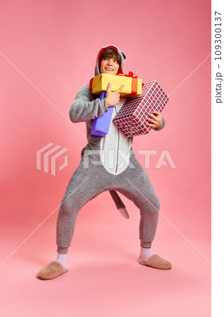 Full length portrait of charming young man standing and thinking about who should be first to give big gift from those that he holds so tightly against pink background Full length portrait of charming young man standing and thinking about who should be first to give big gift from those that he holds so tightly against pink background 109300137