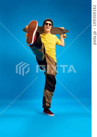 Full length portrait of cool dude rastaman, skateboarder holding skateboard and posing against blue studio background. 109300162