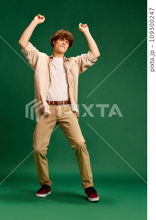 Full length portrait of young man, student dressed smart casual retro outfit dancing raising hands of joy against green studio background. Full length portrait of young man, student dressed smart casual retro outfit dancing raising hands of joy against green studio background. 109300247