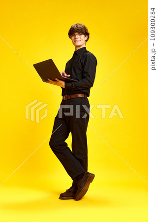 Study and work online from anywhere in world. Full length portrait of young attractive man posing with laptop against vivid yellow studio background. 109300284