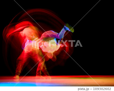 Young athletic man, b-boy dancing hip-hop, breakdance on one hand raising legs up against black background in neon light with motion blur Young athletic man, b-boy dancing hip-hop, breakdance on one hand raising legs up against black background in neon light with motion blur 109302602