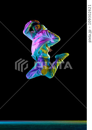 Studio shot of young flexible sportive man dancing breakdance, jumping, freezing in casual outfit against black background in neon light 109302621