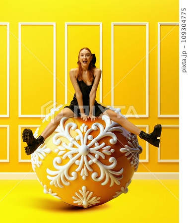 Happy, excited, emotional young girl in black dress sitting on big Christmas tree decoration, toy over bright yellow background. Christmas and New Years holidays Happy, excited, emotional young girl in black dress sitting on big Christmas tree decoration, toy over bright yellow background. Christmas and New Years holidays 109304775