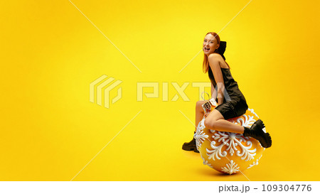 Happy smiling young girl in elegant dress sitting on giant Christmas tree toy, decoration over bright yellow background. Christmas and New Year 109304776
