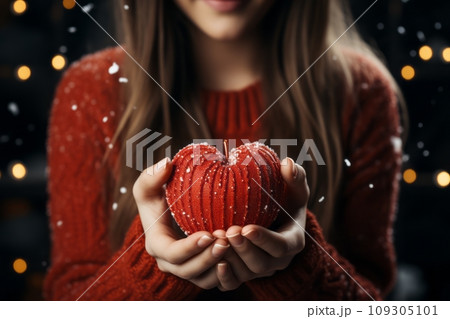 A young woman with long, curly hair, wearing a red sweater. She is holding a red, knitted Christmas heart in her hands. A young woman with long, curly hair, wearing a red sweater. She is holding a red, knitted Christmas heart in her hands. 109305101