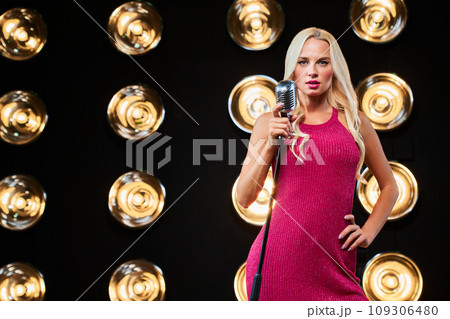 Beautiful female pop star singer performing music with retro microphone on stage. Front view of slim blonde lady posing with mic stand, in front of bulb lights wall on background. Concept of perform. 109306480
