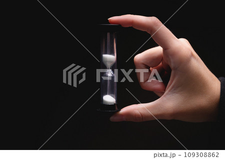 Close-up of a woman holding an hourglass in the dark. Fingers hold on both sides a plastic sand clock with white sand on a black background Close-up of a woman holding an hourglass in the dark. Fingers hold on both sides a plastic sand clock with white sand on a black background 109308862