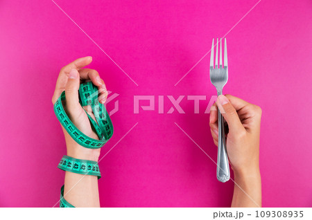 Close-up of a hand with a measuring tape wrapped around it and a fork in the other hand. Diet and healthy eating concept Close-up of a hand with a measuring tape wrapped around it and a fork in the other hand. Diet and healthy eating concept 109308935