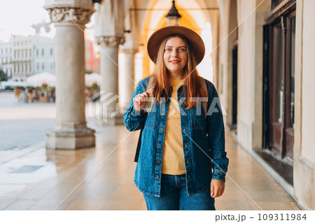 Attractive young female tourist is exploring new city. Redhead girl in hat posing on Market Square in Krakow. Traveling Europe in autumn. High quality photo, The Cloth Hall, Vacation concept 109311984