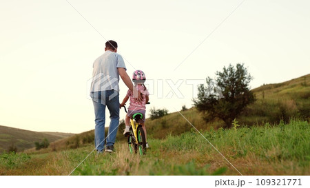 Attentive father teaches preschooler girl to ride bicycle in sunset field 109321771