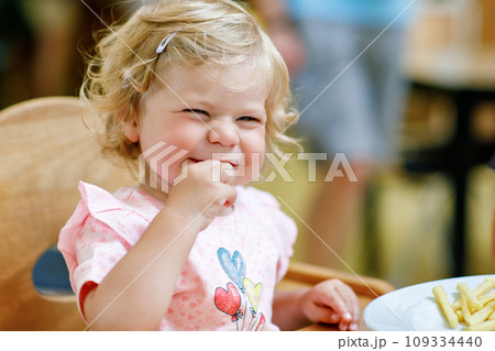 Adorable toddler girl eating healthy vegetables and unhealthy french fries potatoes. Cute happy baby child taking food from dish at daycare or nursery canteen. 109334440
