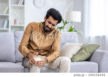 Indian young man sitting on the sofa at home and holding his knee, suffering and grimacing in pain. 109336456