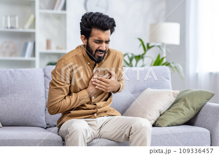 A young Indian man is sitting on the couch at home and holding his hands to his chest, feeling a strong pain in his heart, breathing hard. A young Indian man is sitting on the couch at home and holding his hands to his chest, feeling a strong pain in his heart, breathing hard. 109336457