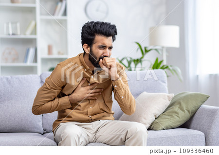 Indian young man sitting at home on the couch and coughing, covering his mouth, holding his chest, feeling severe pain, suffering from an infectious disease. 109336460