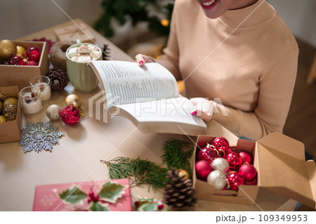 Young asian woman in sweater reading a book with happiness and drinking hot chocolate with marshmallow after decorating ornaments for celebrate christmas festive holiday and winter seasons at home 109349953