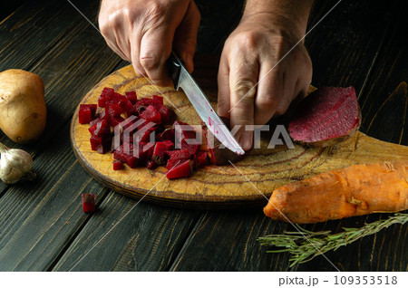 Cutting boiled beets into small pieces with a knife to prepare a vinaigrette. Knife in the hand of a cook on the kitchen table. Vegetable dish concept. Cutting boiled beets into small pieces with a knife to prepare a vinaigrette. Knife in the hand of a cook on the kitchen table. Vegetable dish concept. 109353518
