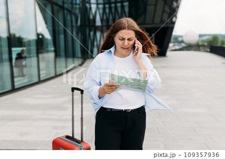 Unhappy 30s woman tourist dressed in casual look speaking on smartphone gadget on the city street. Angry redhead girl with a red suitcase and map standing on city street. Negative people emotion. Unhappy 30s woman tourist dressed in casual look speaking on smartphone gadget on the city street. Angry redhead girl with a red suitcase and map standing on city street. Negative people emotion. 109357936