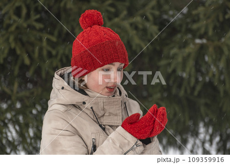 Close-up portrait of girl in stylish red hat with mittens on spruce branches background. Middle-aged woman in comfortable winter clothes 109359916