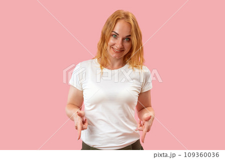 Smiling woman points her fingers down on pink background looks in camera. Portrait of young smiling round-faced woman with ginger hair. 109360036
