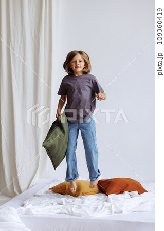 A child jumping on the bed with a pillow in his hands against the background of a white wall A child jumping on the bed with a pillow in his hands against the background of a white wall 109360419