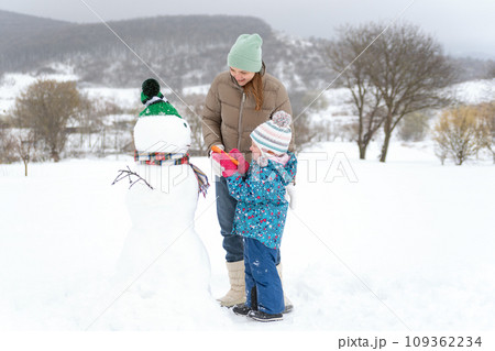Cute toddler and mon playing with snowman outdoors in winter. Winter weekend with family in the countryside. Cute toddler and mon playing with snowman outdoors in winter. Winter weekend with family in the countryside. 109362234