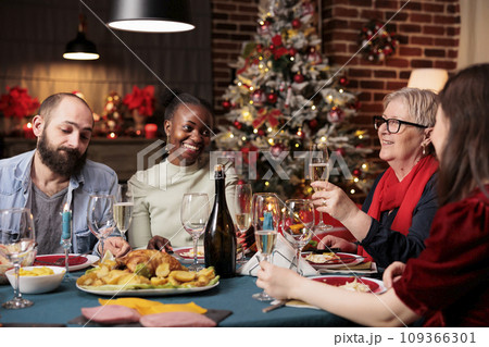 Grandma makes toast at dinner table with friends and family, enjoying christmas eve festivity at home. Senior woman giving speech with raised wine glasses for cheers, winter celebration. 109366301
