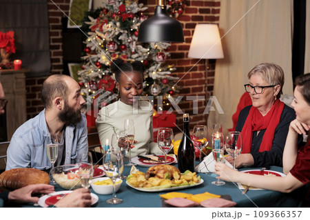People raising glasses for toast during christmas dinner, enjoying family time at home with traditional food. Diverse friends making speech to thank everyone for celebrating around the table. 109366357